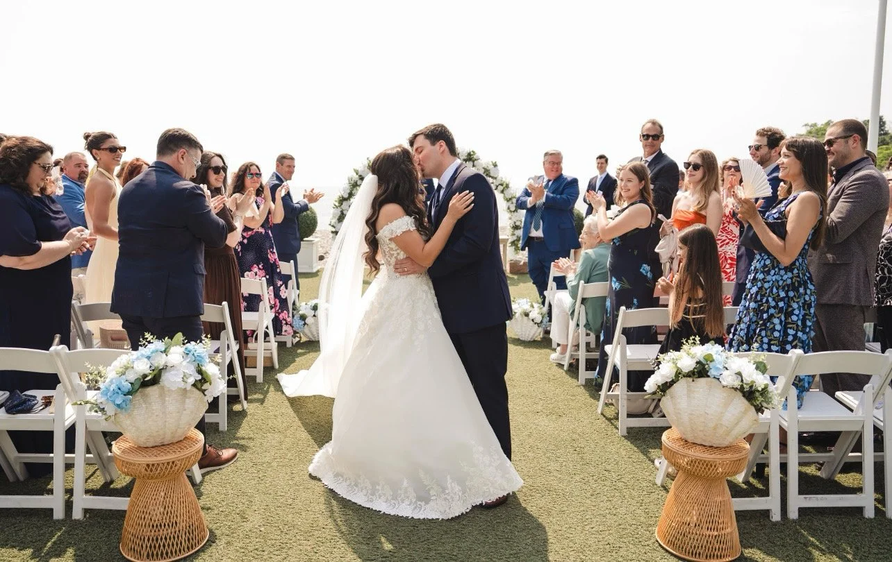 A bride and groom kiss during their outdoor wedding ceremony surrounded by seated and standing guests clapping, with white chairs, floral arrangements, and a sunny sky in the background.