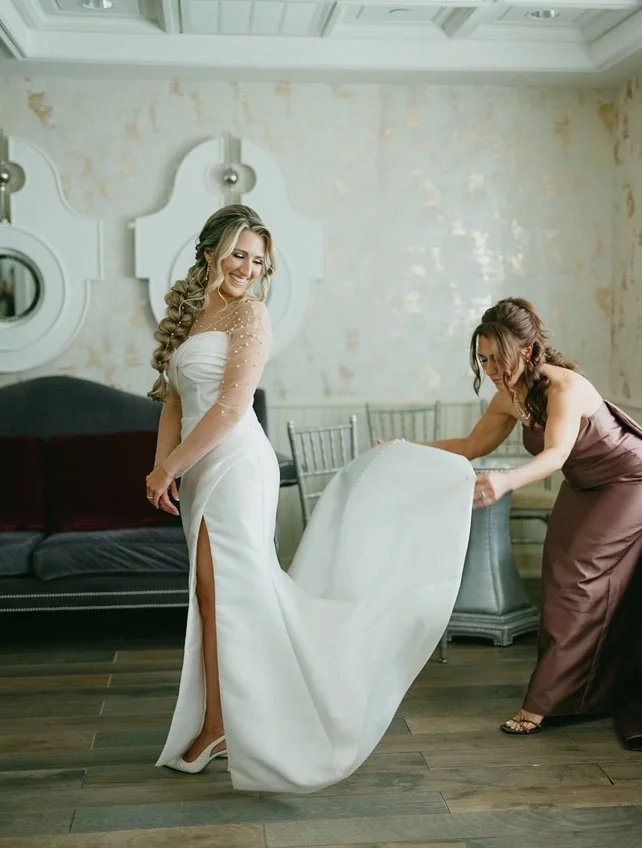 A woman in a white wedding dress is smiling as she stands while another woman assists with her dress in a room with a distressed wall and wooden floor.
