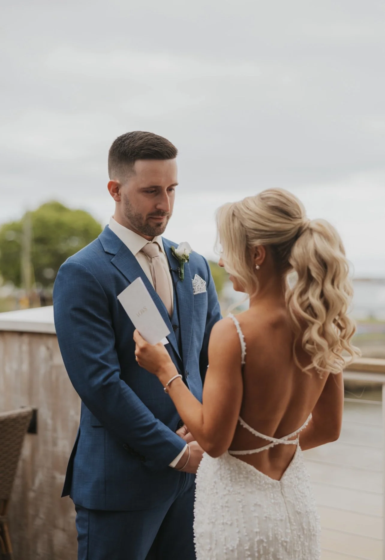 A bride and groom during their wedding ceremony outdoors. The bride, with long curly blonde hair, is holding a marriage certificate and looking at the groom, who is dressed in a blue suit with a boutonniere.