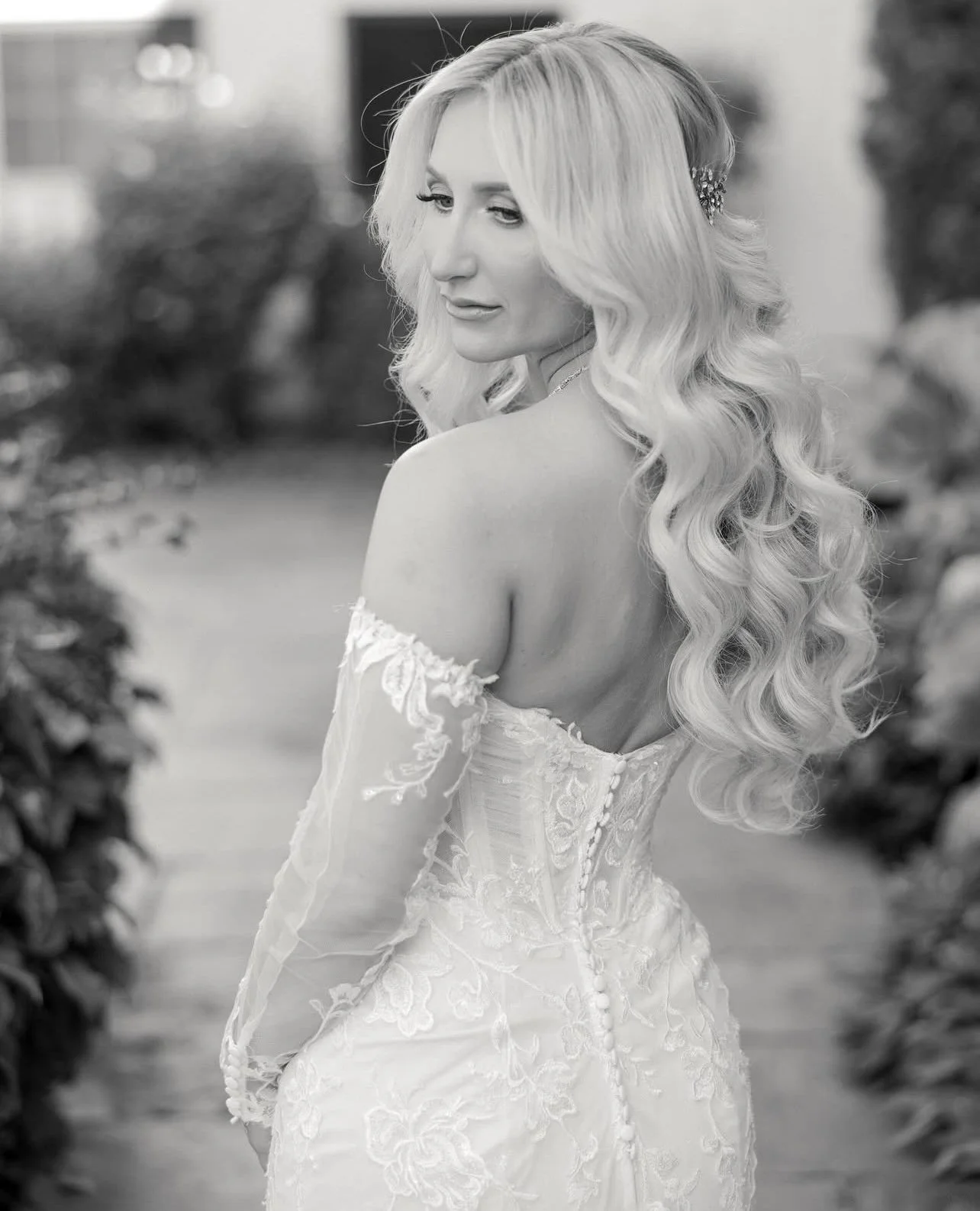 Black and white photo of a woman with long, wavy blonde hair, wearing a lace dress, standing outdoors with blurred greenery in the background.