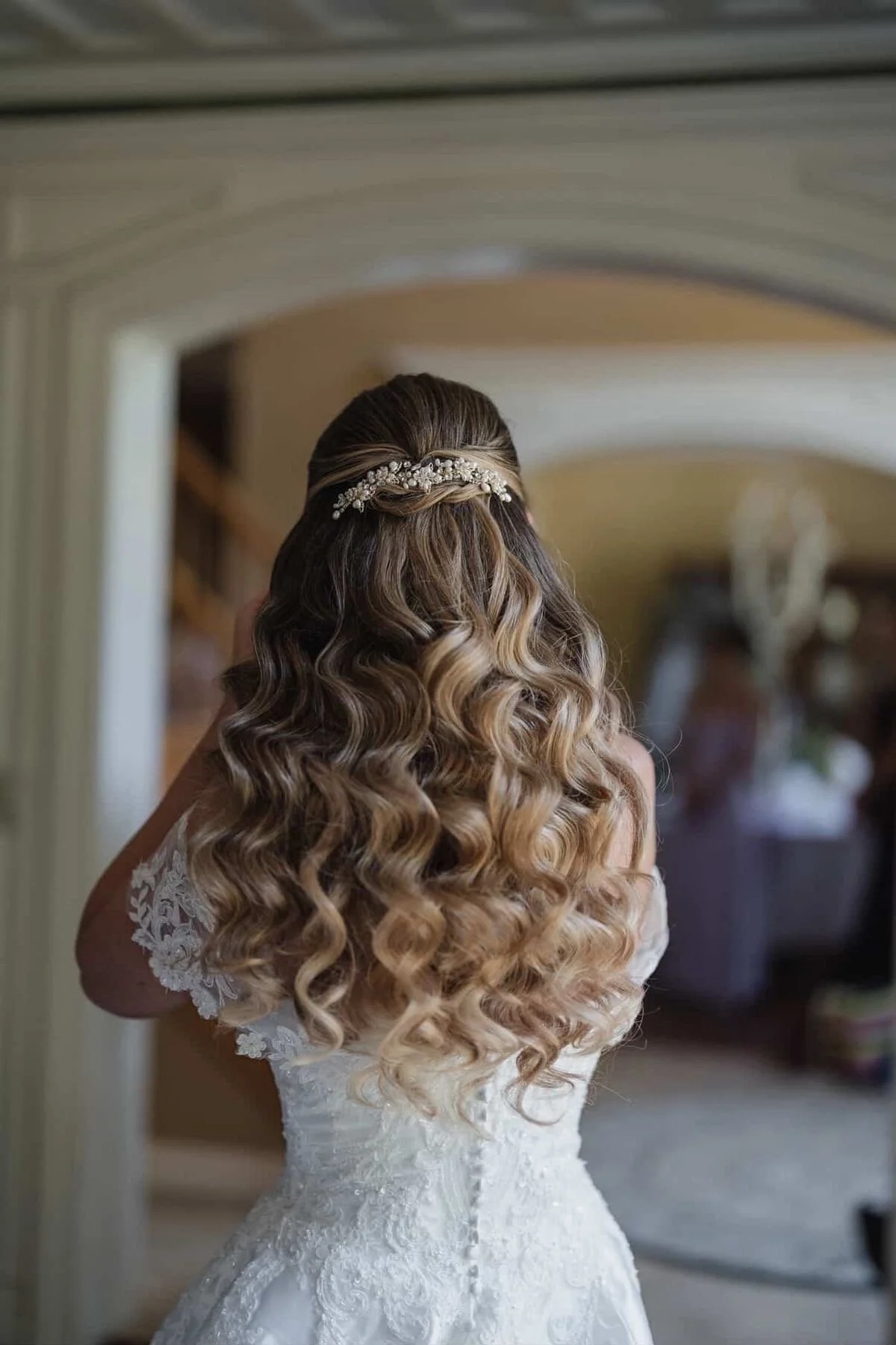 Back view of a woman with long, curly hair wearing a white dress and a decorative hairpiece, standing in front of a mirror.