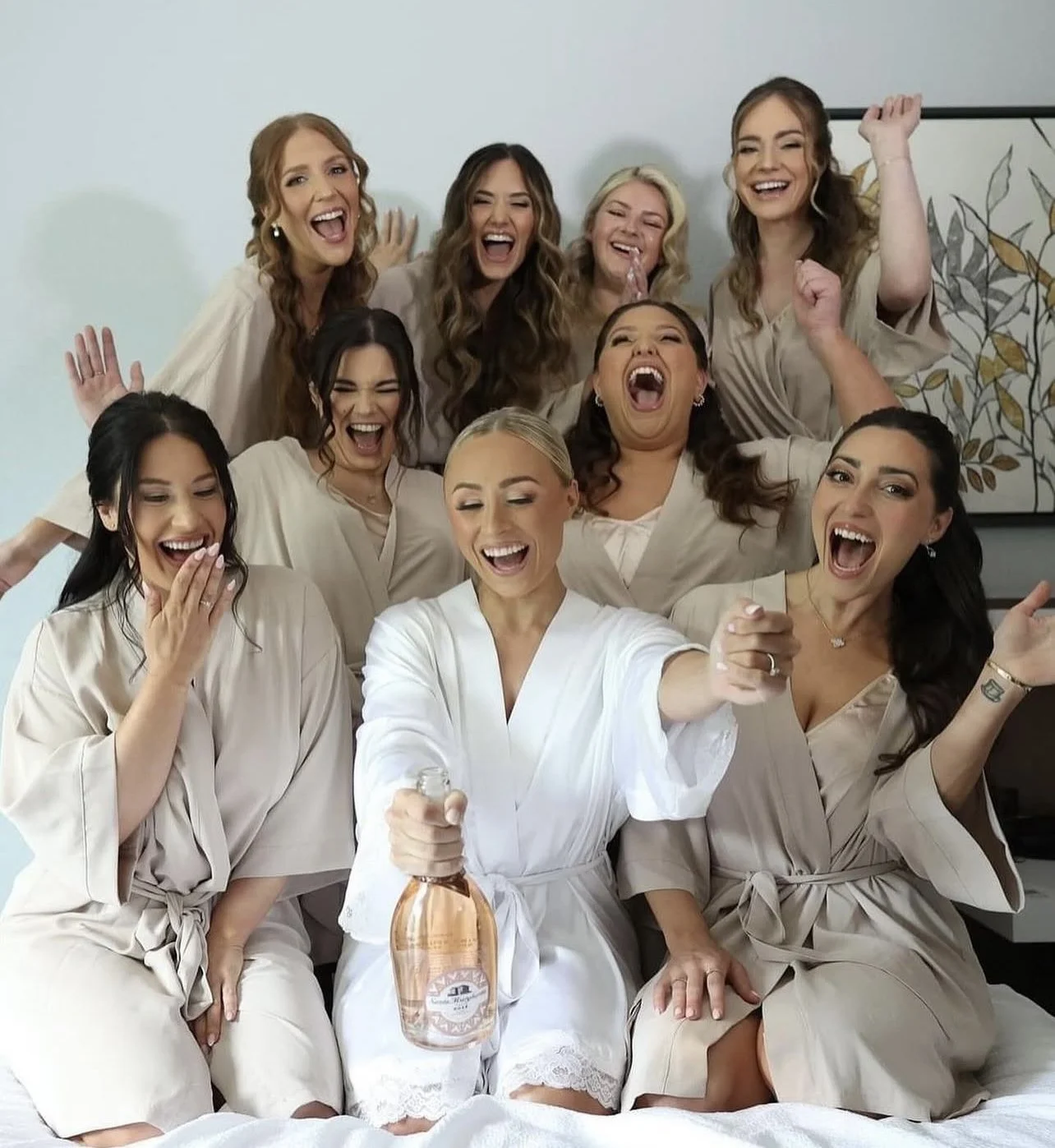A group of women in cream-colored robes celebrating and smiling, with one woman in white holding a bottle of champagne.