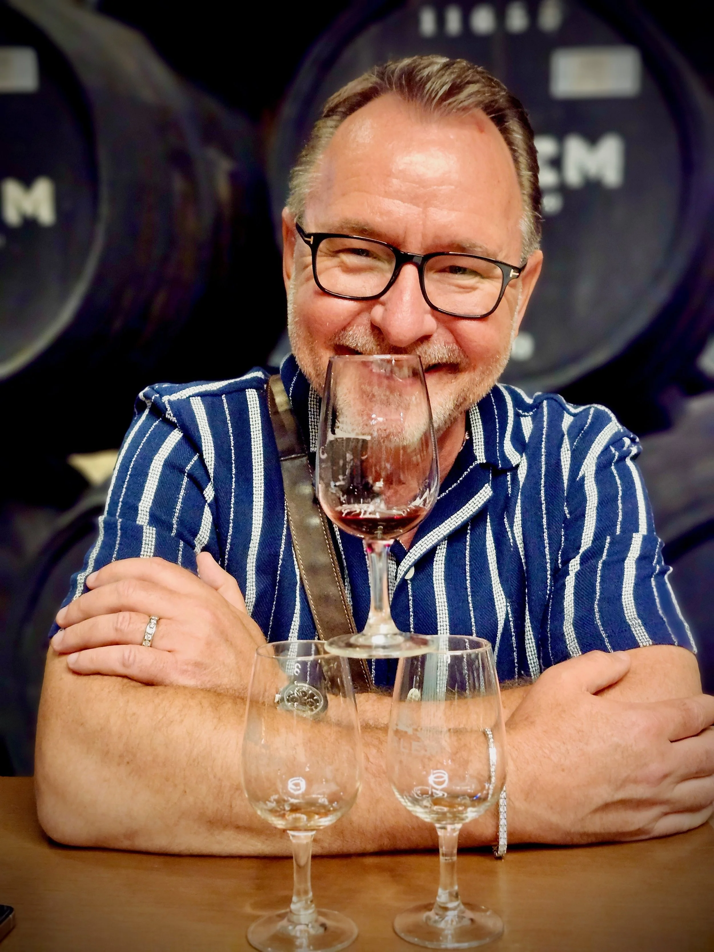 A smiling man with glasses, wearing a striped blue shirt, sitting at a table with a glass of red wine, with wine barrels in the background.