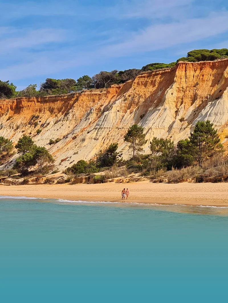 Two people walking on a sandy beach with cliffs and trees in the background, under a blue sky with some clouds.