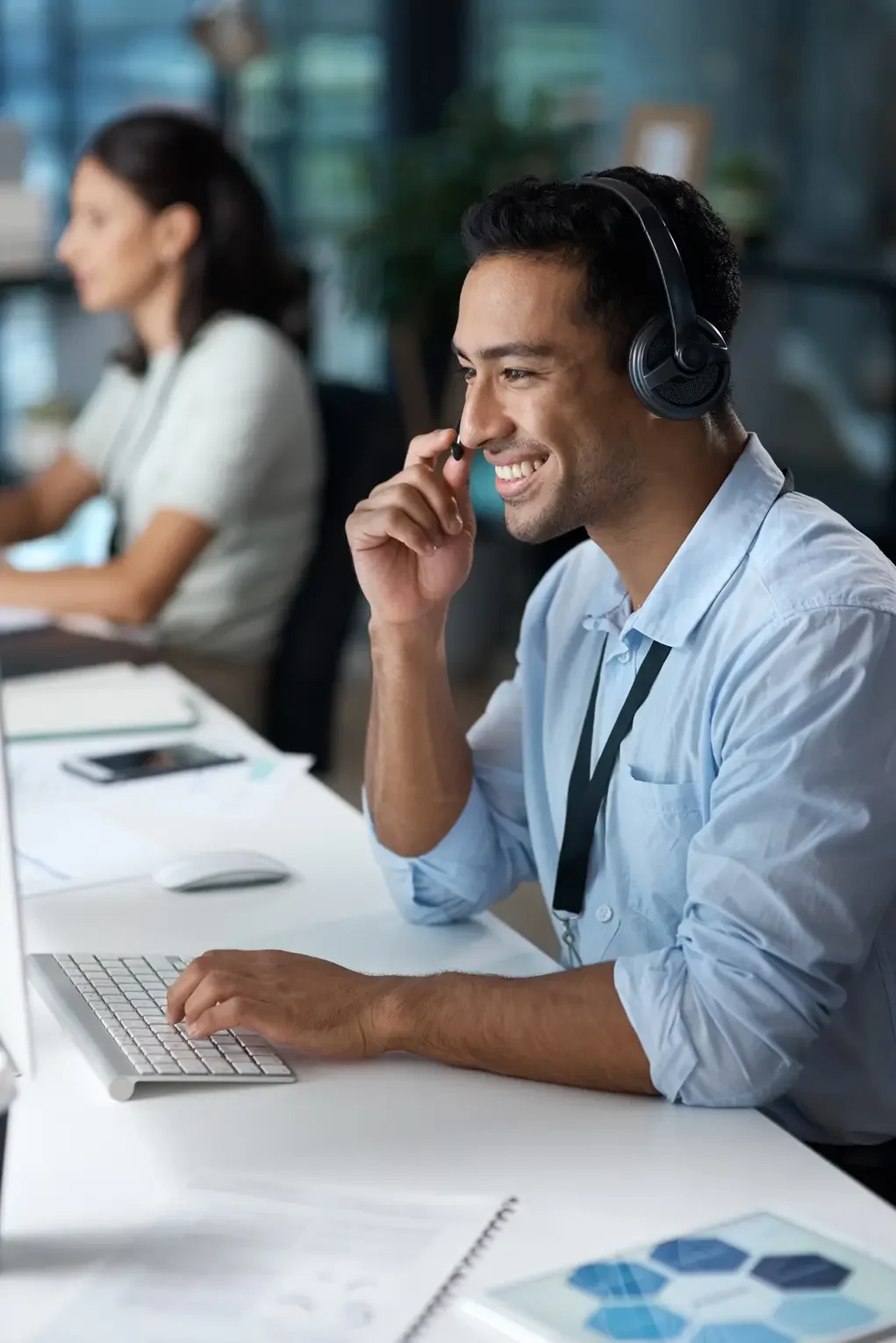 A smiling man wearing a headset and a light blue shirt working at a desk with a computer, paper, and a smartphone in an office environment.