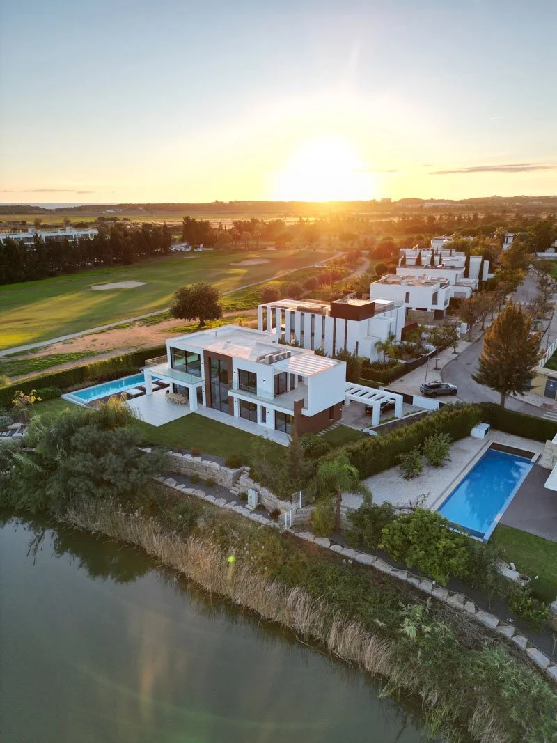 Aerial view of modern white houses with swimming pools near a golf course and waterway at sunset.