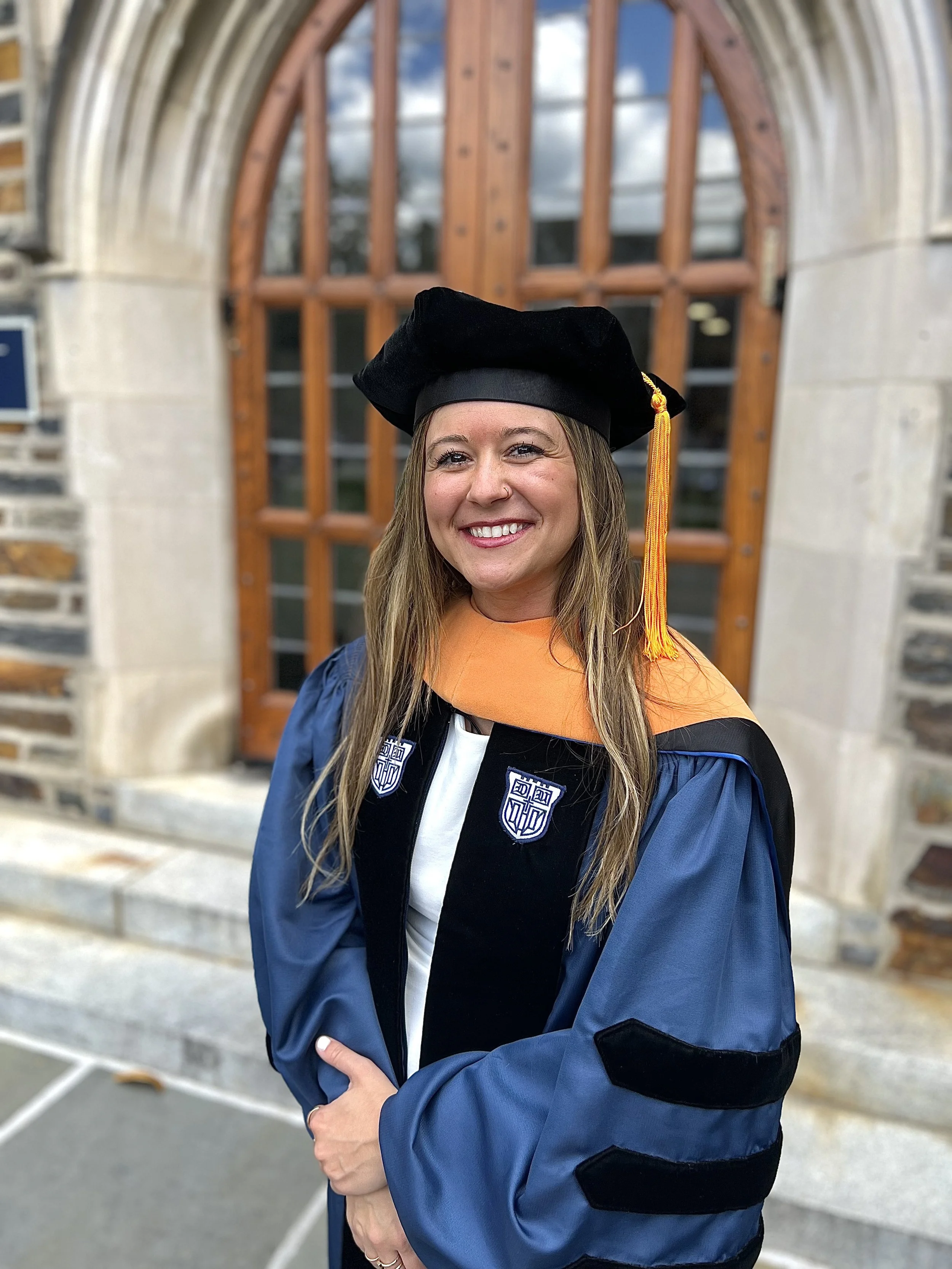 A woman in graduation regalia, including a black cap and gown with blue sleeves, standing in front of a wooden door with stone accents and smiling.