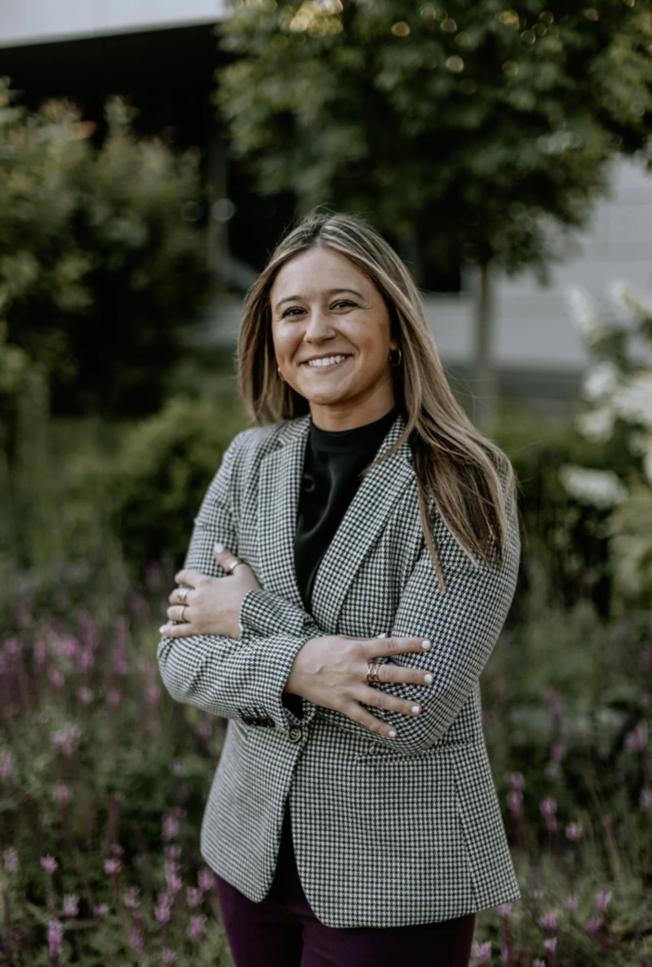 A woman with shoulder-length light brown hair, smiling, wearing a checkered blazer over a black top, standing outdoors in a garden with green trees and pink flowers in the background.