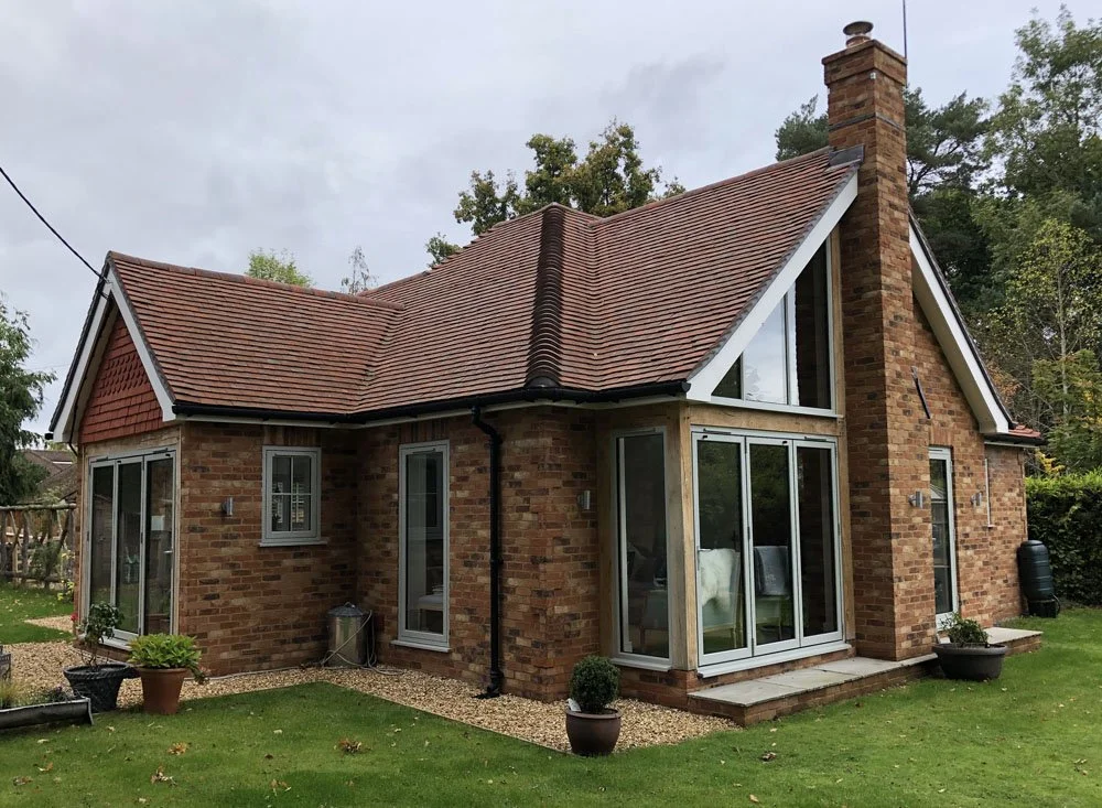 A brick house with a red tiled roof, large windows, and a prominent chimney, surrounded by a lawn and trees.