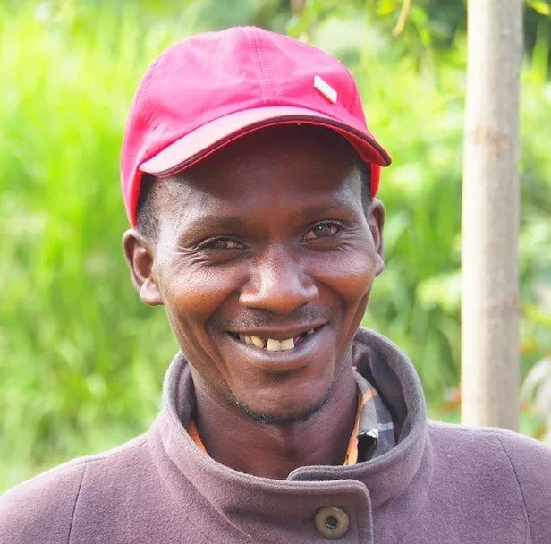 Ein Mann mit dunkler Haut trägt eine rote Kappe und einen grauen Mantel. Er lächelt und hat große Zähne. Im Hintergrund sind grüne Pflanzen.