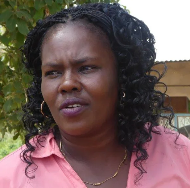 Eine schwarze Frau mit lockigem Haar trägt ein pinkfarbenes Hemd und eine goldene Halskette. Sie steht im Freien vor Bäumen und einem Holzgebäude, sieht nachdenklich aus.