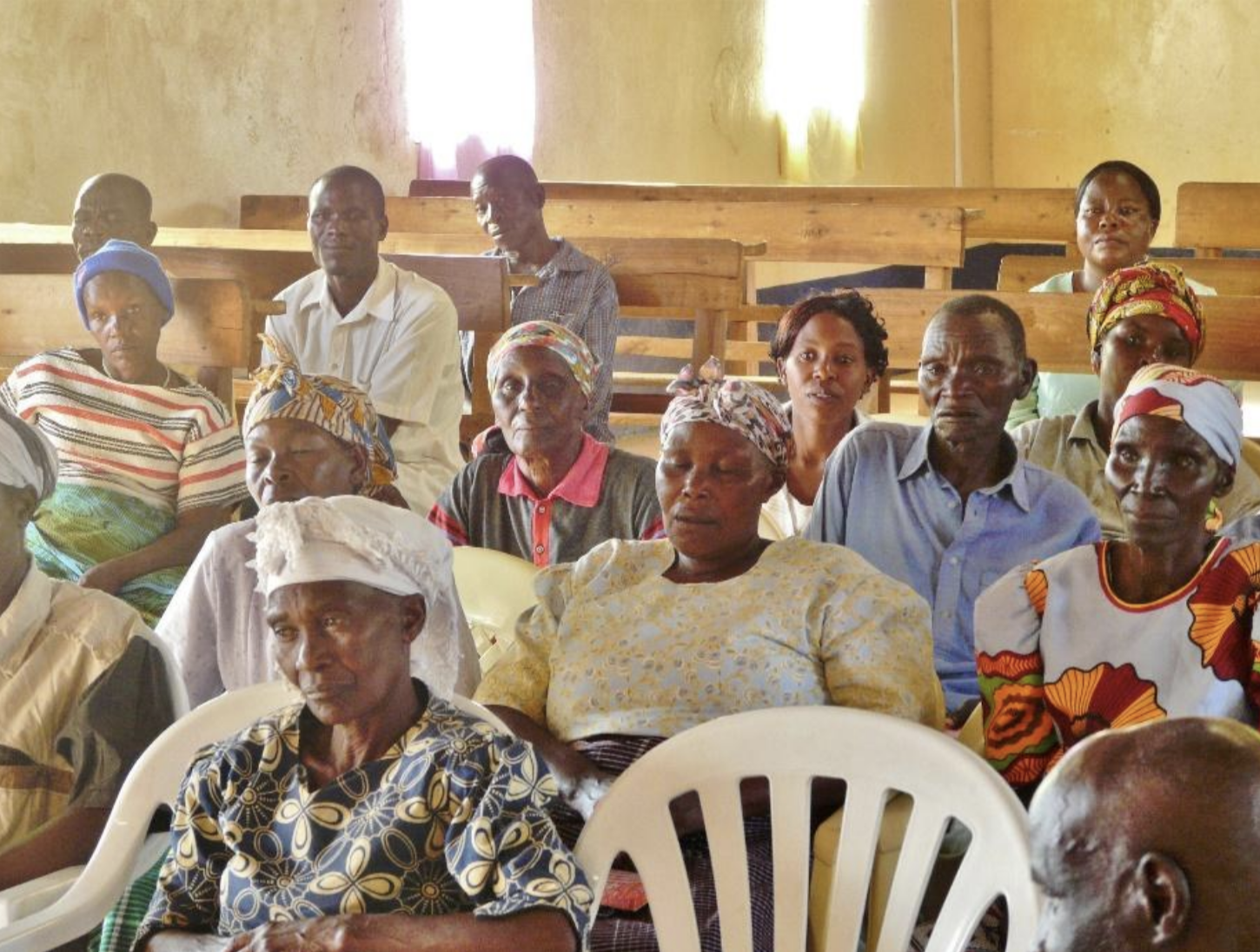 Menschen sitzen in einem Raum auf Bänken und Stühlen, einige tragen traditionelle Kleidung und Kopftücher, in einfachen, gelben Wänden, wahrscheinlich bei einer Gemeinschaftsversammlung.