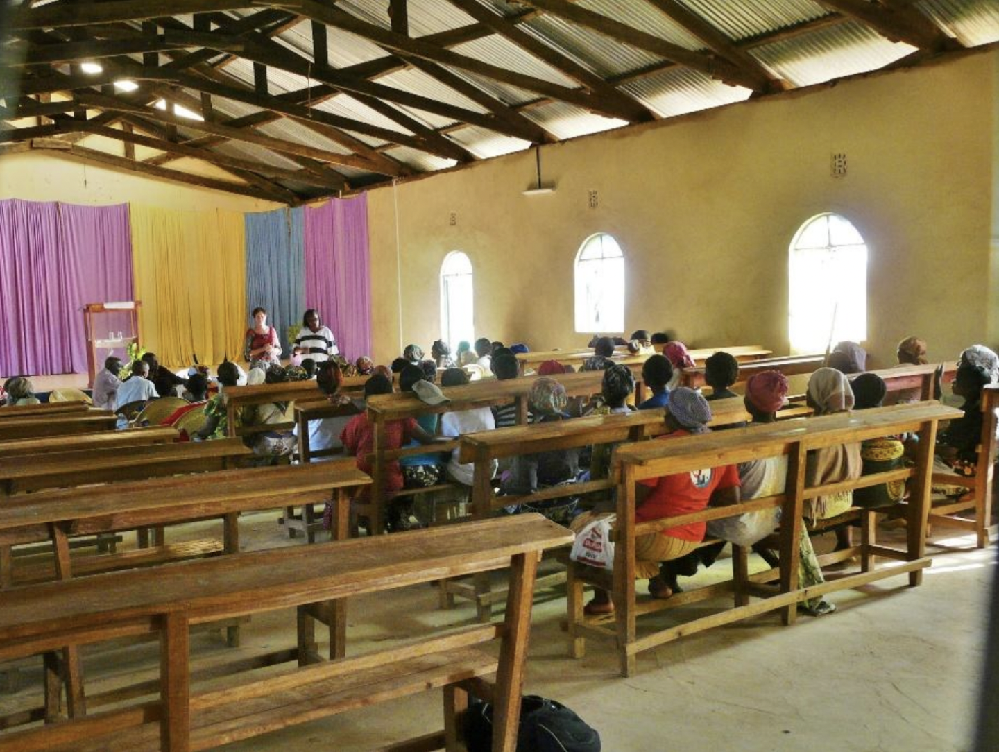 Menschen sitzen in einer Kirche oder Gemeinschaftsraum mit Holzbänken, vor einem bunten Kulissenhintergrund, während zwei Frauen vorne sprechen.
