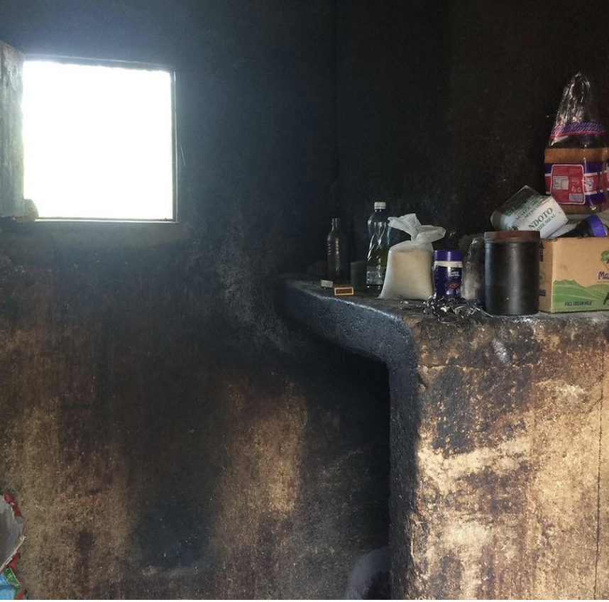 Ein dunkler Raum mit einer kleinen Fensterscheibe. Auf einer schwarzen, verbrannten Oberfläche stehen diverse Hygieneprodukte, Wasserflaschen und eine Teekanne.