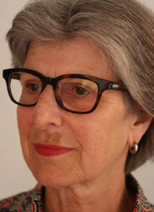 Nahaufnahme einer älteren Frau mit kurzgrauem Haar, schwarzen Brillen und rotem Lippenstift, lächelnd.