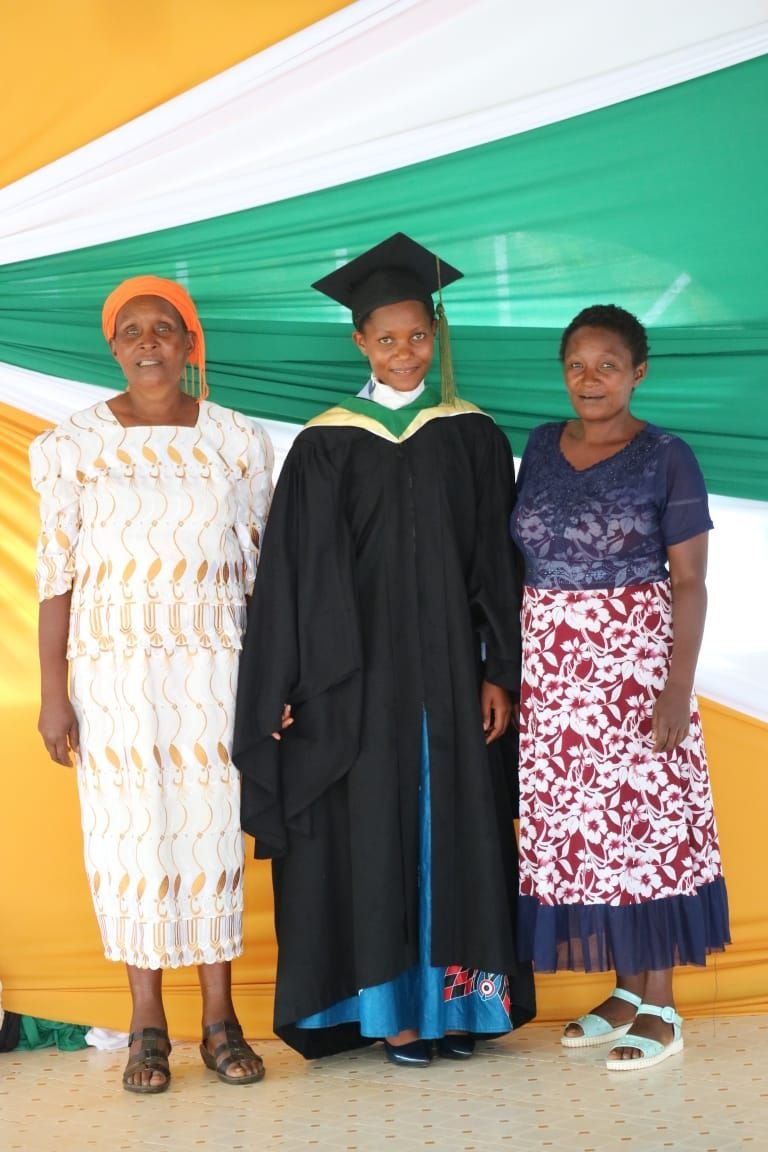 Drei Frauen und eine junge Frau in einem schwarzen Graduation-Gewand und Abschlussmütze bei einer Abschlussfeier, im Hintergrund bunte Stoffdekorationen.