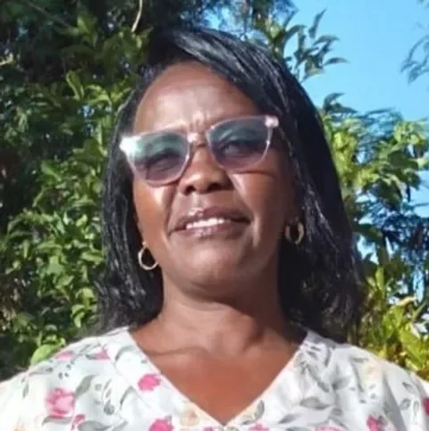 Eine Frau trägt Sonnenbrille und ein weißes T-Shirt mit Blumenmuster, steht vor Bäumen bei sonnigem Himmel.