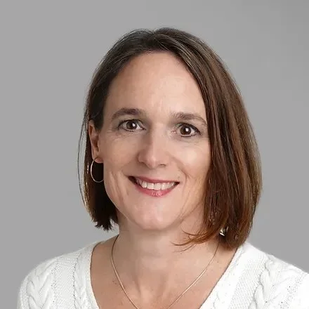 Porträt einer lächelnden Frau mit braunen Haaren, weißen Top und silbernen Ohrringen vor grauem Hintergrund.