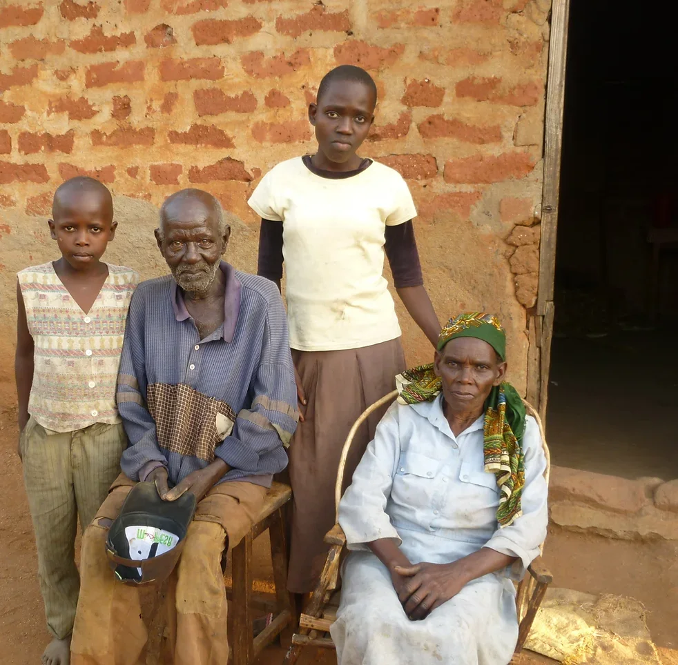 Vier Menschen vor einer Lehmwand, zwei sitzen, zwei stehen. Der Mann im Rollstuhl trägt einen Helm, die Frau sitzt auf einem Stuhl trägt ein grünes Tuch um den Kopf, die Jungen stehen neben ihnen.