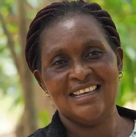 Eine lachende Frau mit dunkler Haut und braunen Haaren, die in Zöpfen geflochten sind, vor einem grünen, natürlichen Hintergrund.