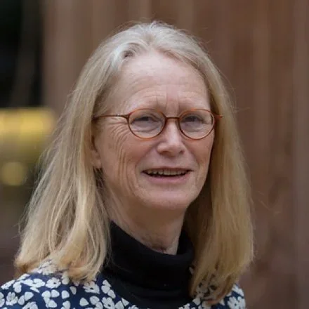 Portrait einer älteren Frau mit langen blondem Haar, runden Brillen und einem schwarzen Tuch, die vor einer Holzwand steht.