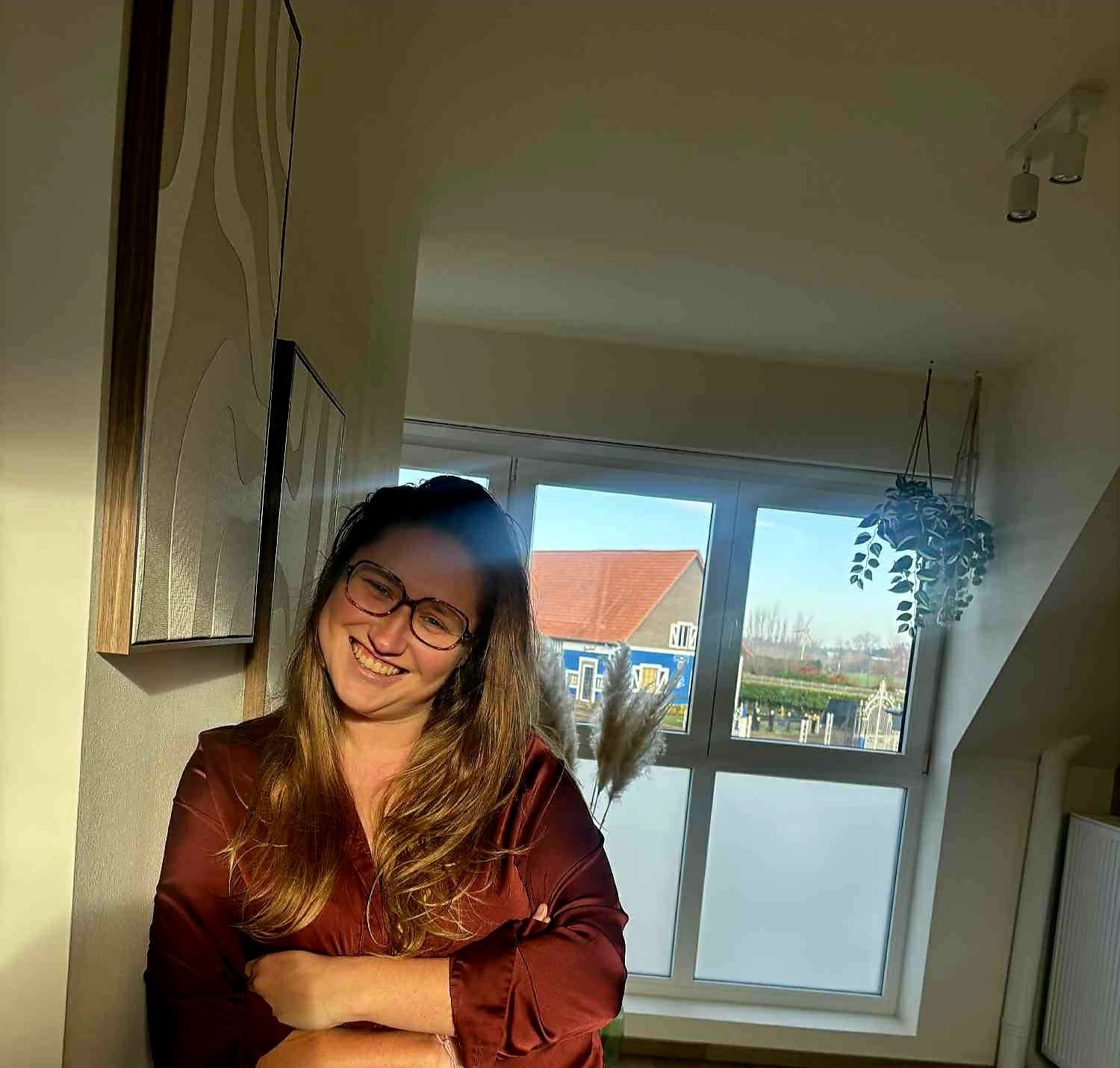 Vrouw met lang haar en brillen, staand bij een witte muur met kunstwerken achter haar, lachend in een lichte kamer met zonnelicht.