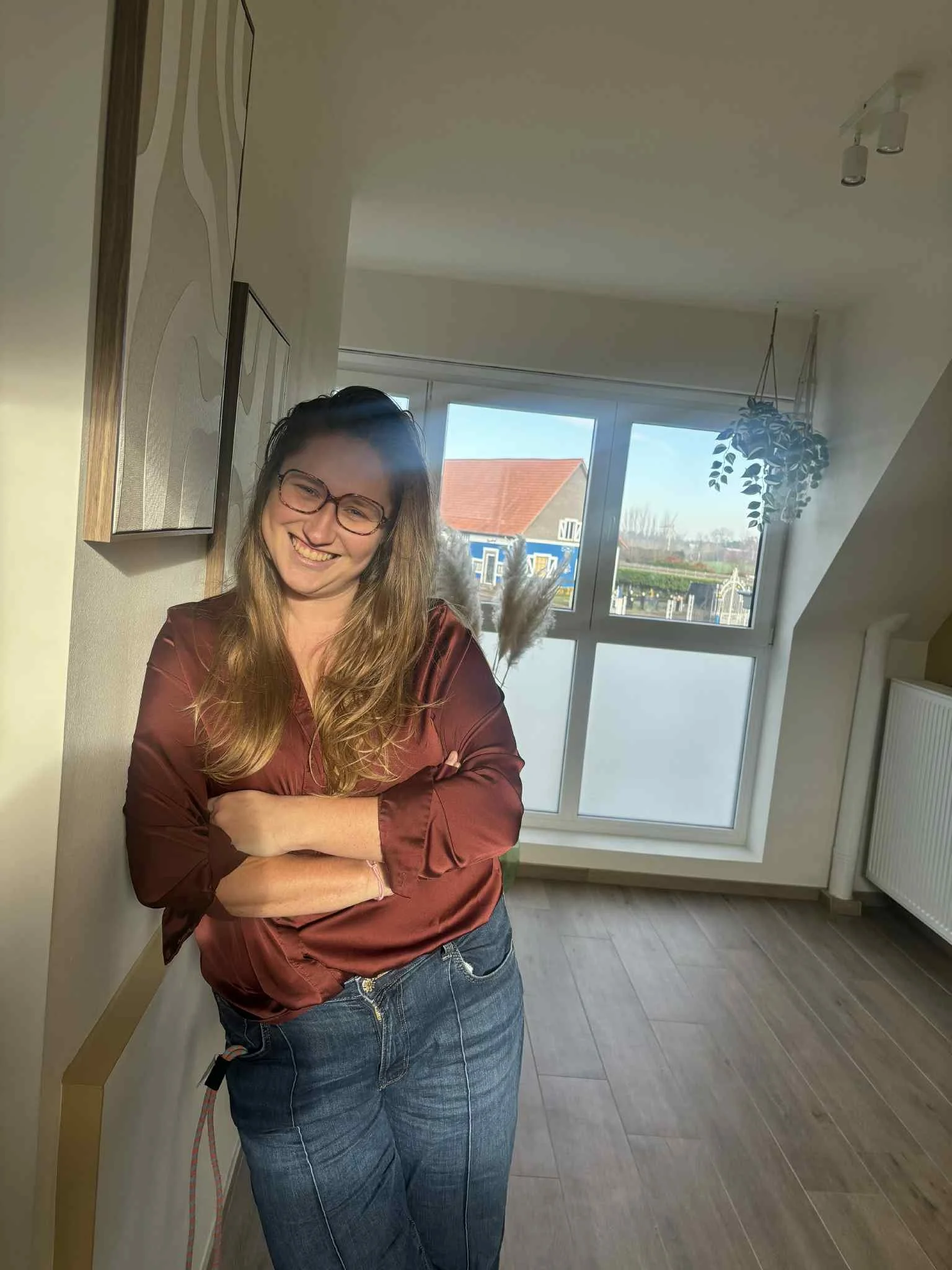 Vrouw met kort haar en brillen zittend aan een witte tafel, lachend. De tafel heeft vazen met gedroogde bloemen, bekers en boeken. De kamer is licht en minimalistisch ingericht.