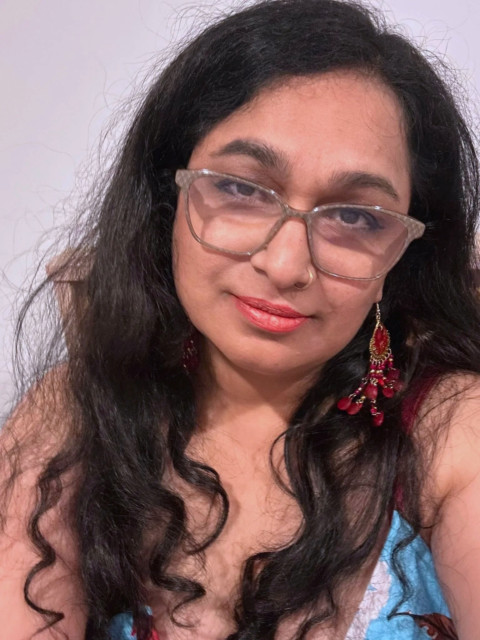 Author is a woman with long, dark, curly hair and she is wearing rose-colored glasses and large red and gold earrings, smiling slightly.