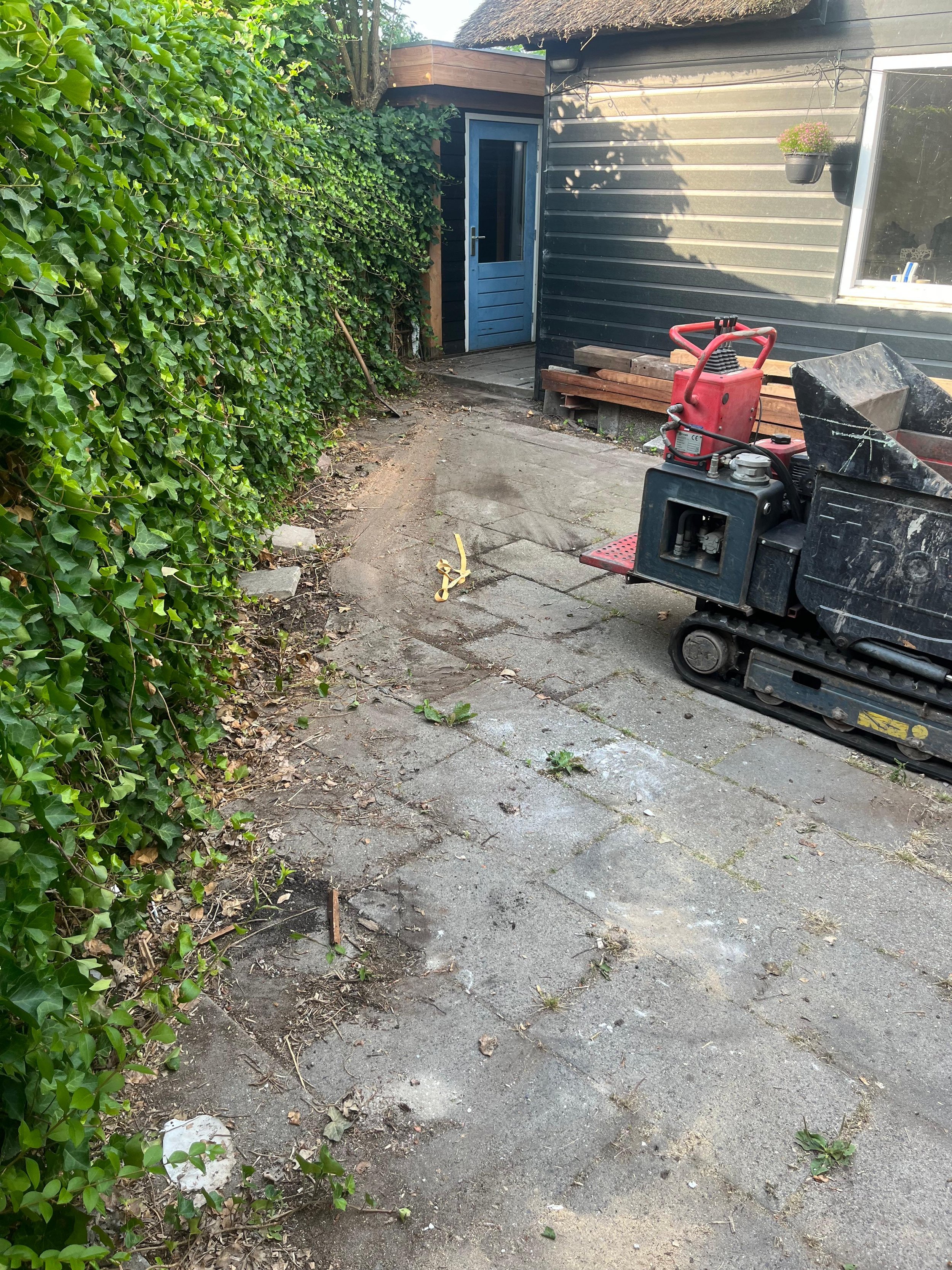A backyard patio with a small construction or landscaping project in progress. There is a tracked mini excavator on the right side, and a pile of wooden planks next to a black house with horizontal siding. A blue door with a window is in the background, and a potted plant hangs on the house wall. The ground has some disturbed dirt and debris, surrounded by green leafy bushes on the left side.