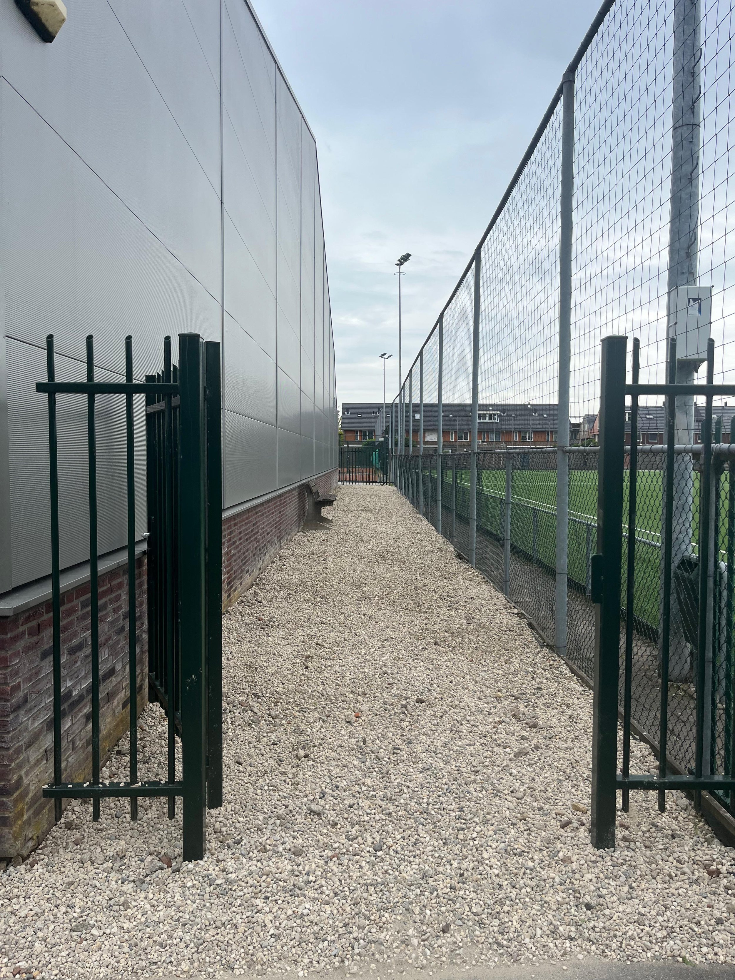 A gravel pathway between a metal building with gray siding and a chain-link fence on a sports field, with a small black gate open on the left.