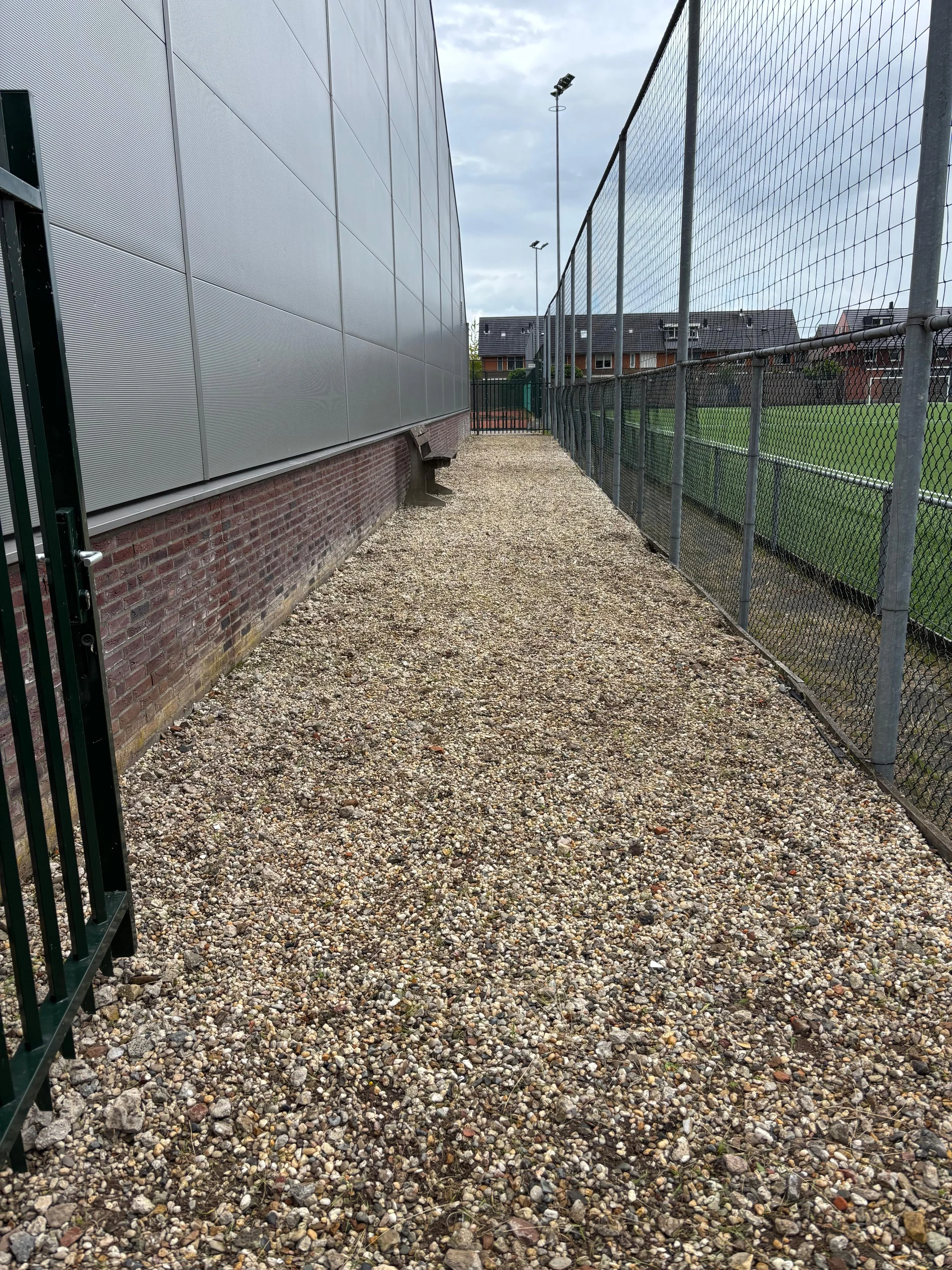 A gravel pathway between a building with a brick base and metallic panels on the left and a chain-link fence with a sports field on the right, under an overcast sky.