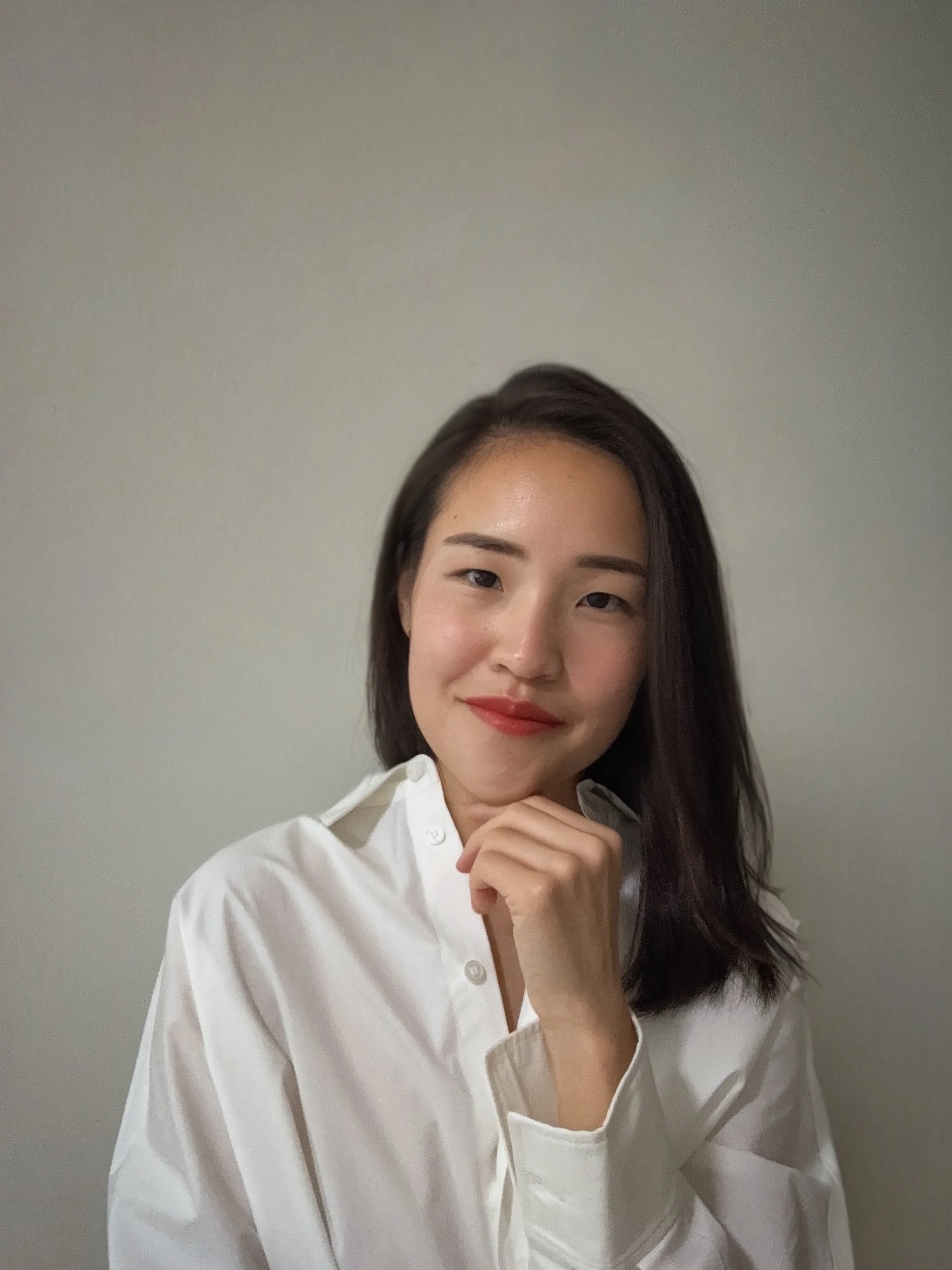 A young woman with dark hair wearing a white shirt, smiling, and resting her chin on her hand, standing against a plain wall.