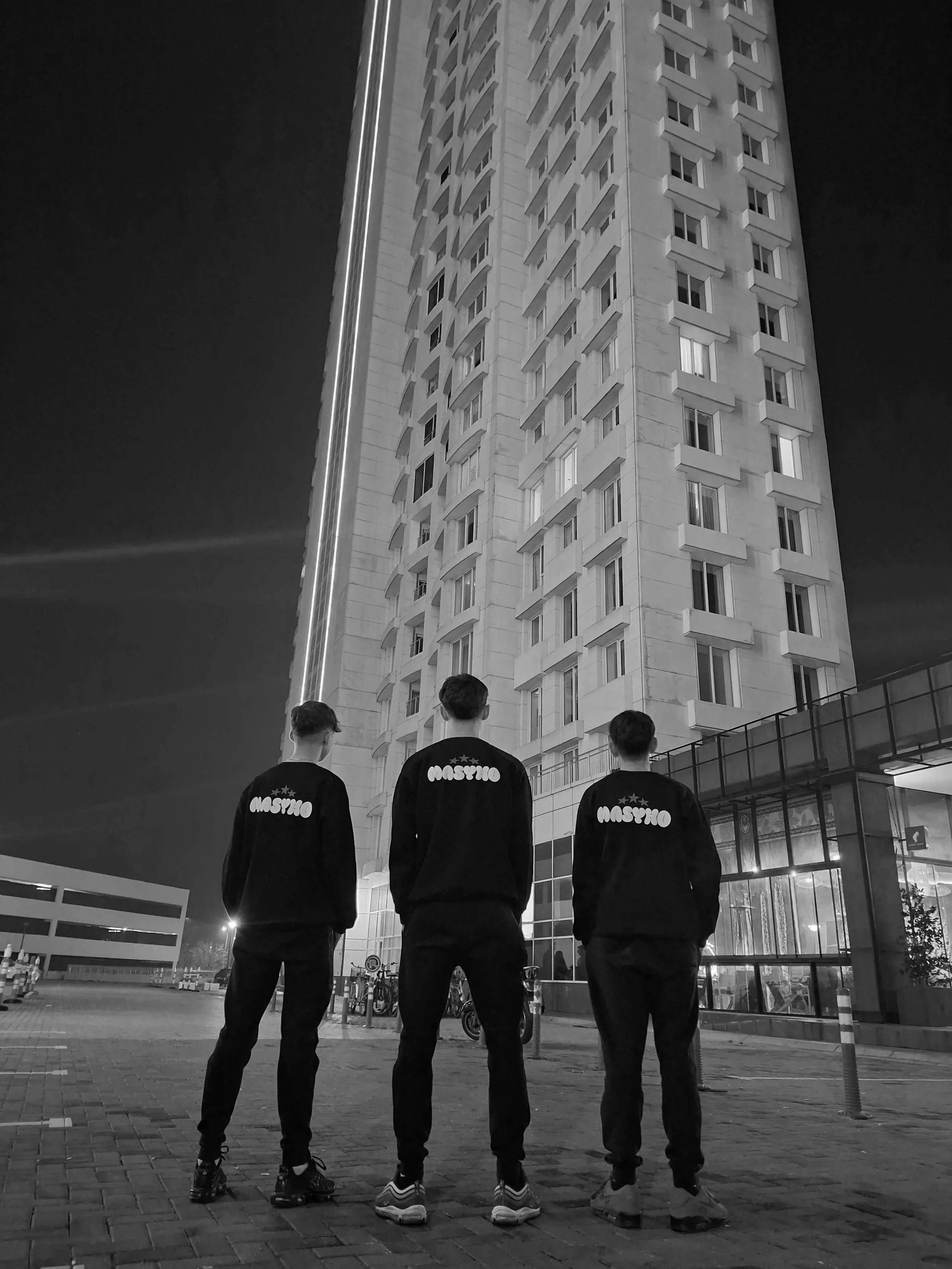 Three young men wearing matching hoodies with the word 'MOSYHO' on back, standing on a city street at night, looking up at a tall modern building.
