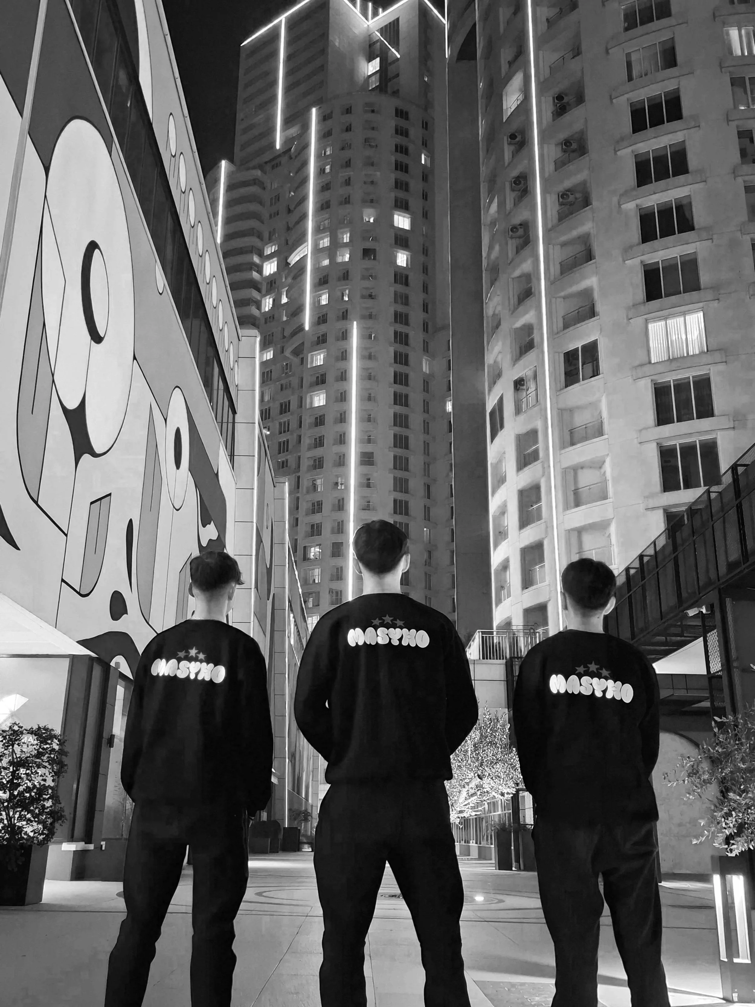 Three young men with short hair, wearing black jackets with the word "Moscow" and stars on the back, standing with their backs to the camera in an urban area at night. They are facing tall buildings with illuminated windows and decorative vertical lights, with one building featuring large wall art, in a cityscape scene.