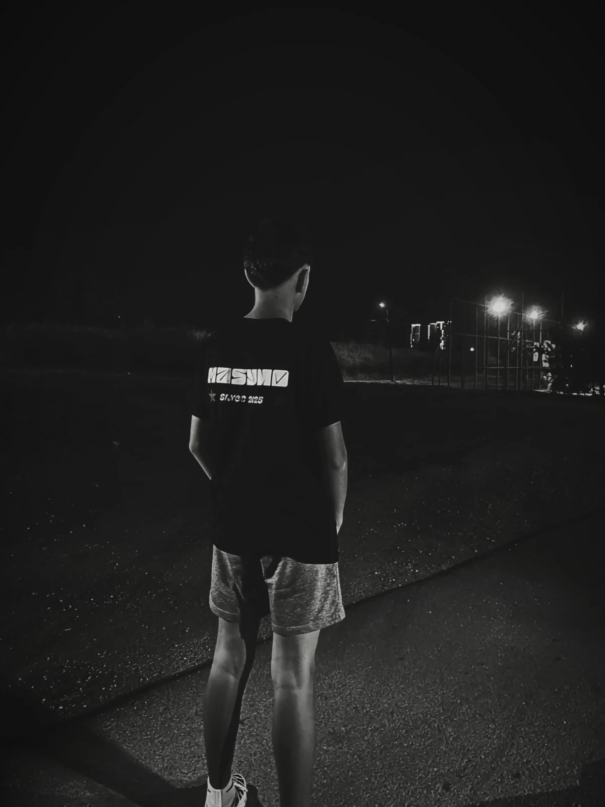 A young person standing outdoors at night, facing away from the camera, wearing a dark MASYHO T-shirt, shorts, and running shoes, with playground equipment and streetlights visible in the background.