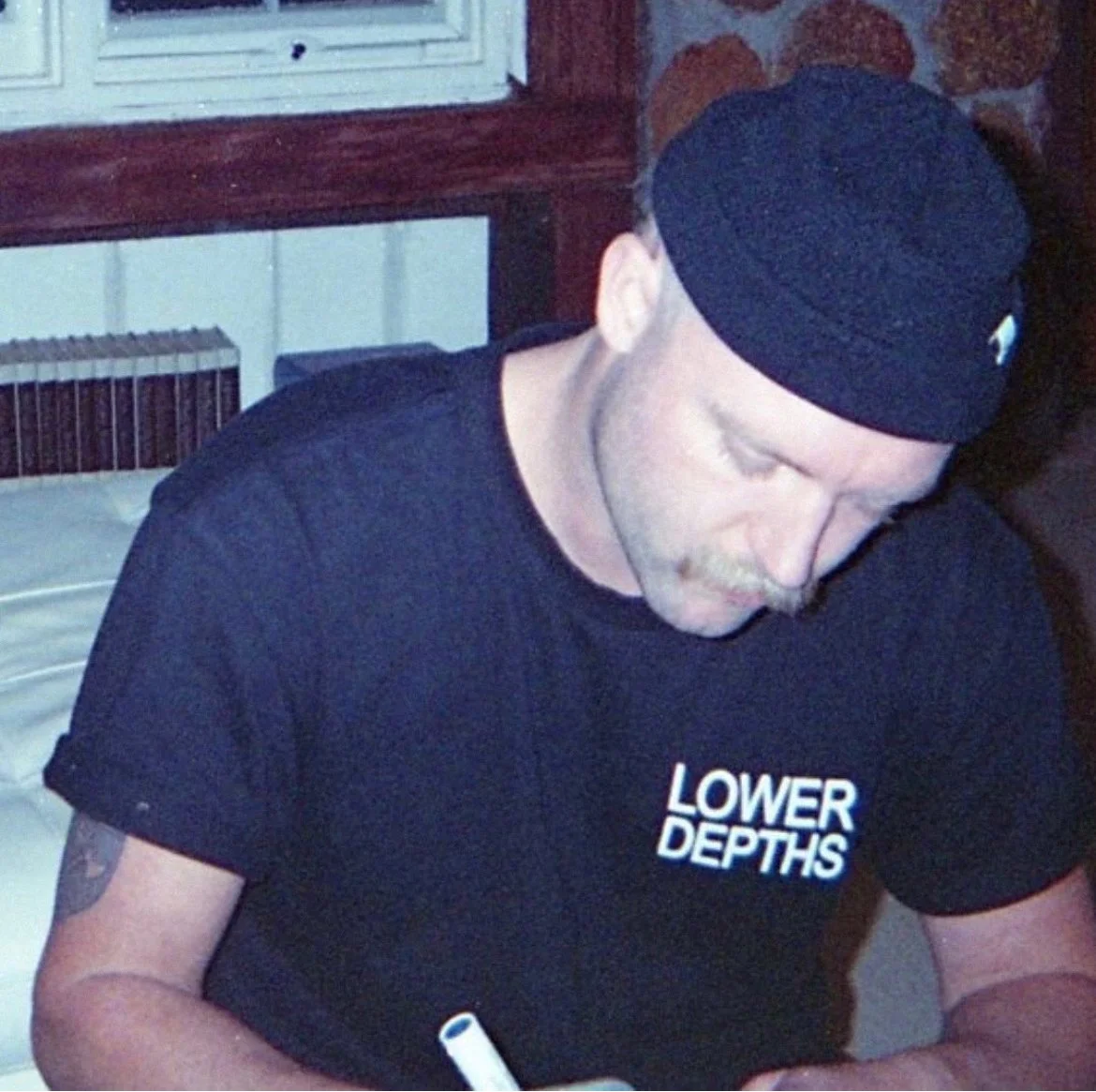 A man with a mustache and a tattoo on his right arm, wearing a black headband and a black T-shirt that says "LOWER DEPTHS," is looking down and writing with a white marker at a table. The background includes a window, radiator, and brick wall.