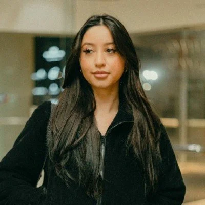 A young woman with long dark hair wearing a black zip-up jacket standing indoors.