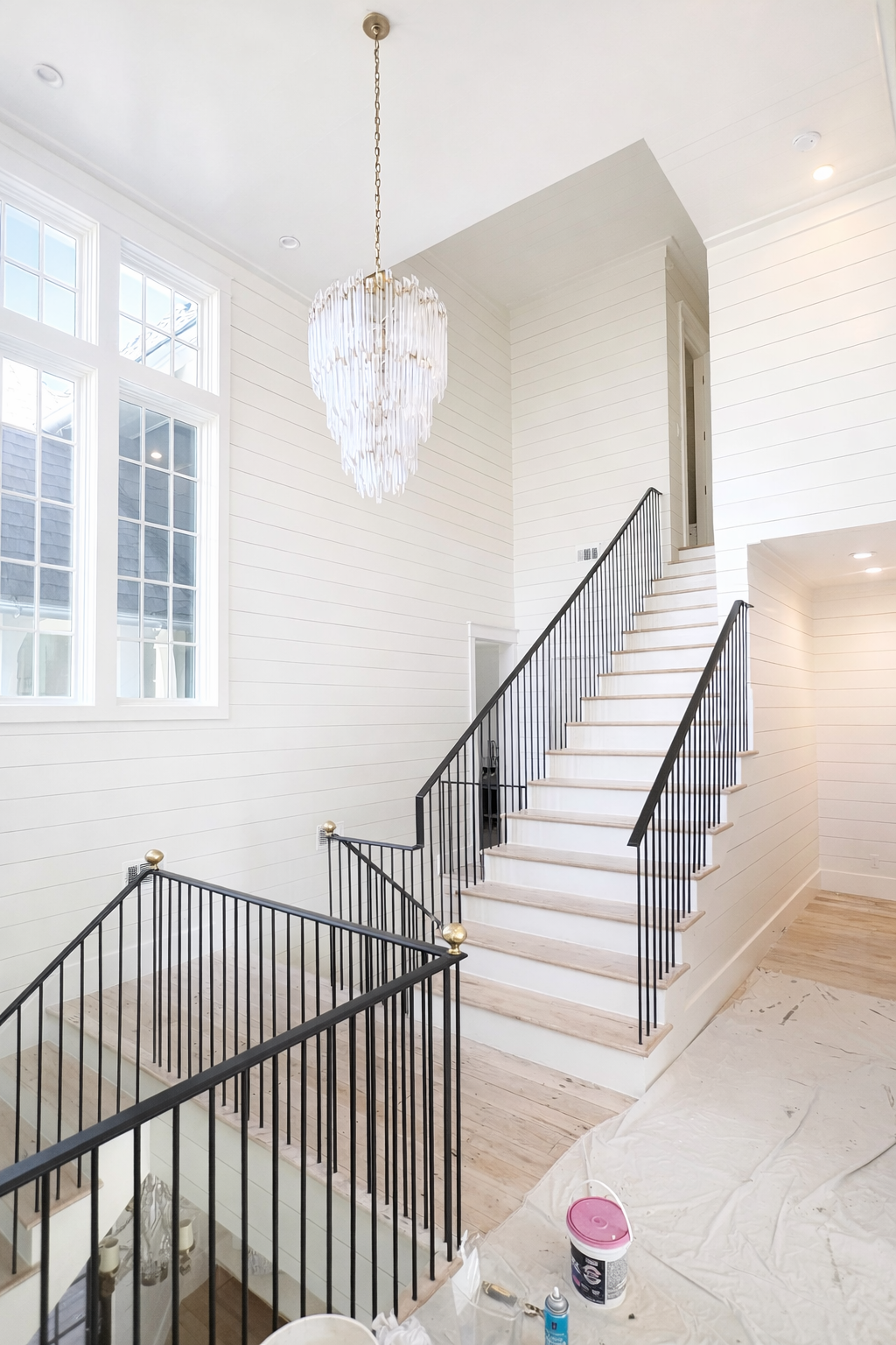 Interior of a modern, bright staircase with white walls, wooden stairs, and black metal railings, with a chandelier hanging from the ceiling and large windows letting in natural light.