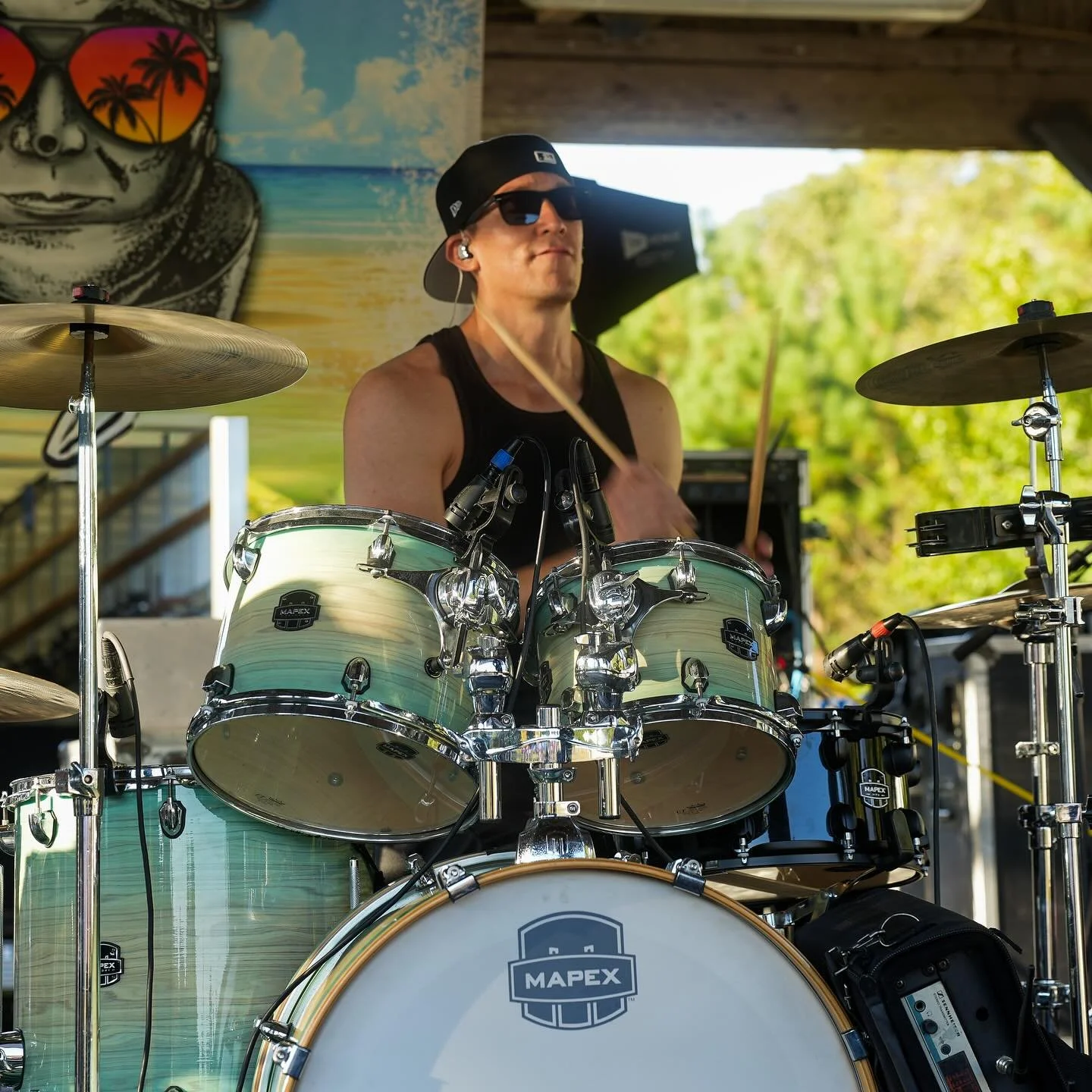 A man wearing sunglasses, a black cap, and a black tank top playing a drum set outdoors. The drum set is beige with a MAPEX logo, and the background has a colorful mural and green trees.