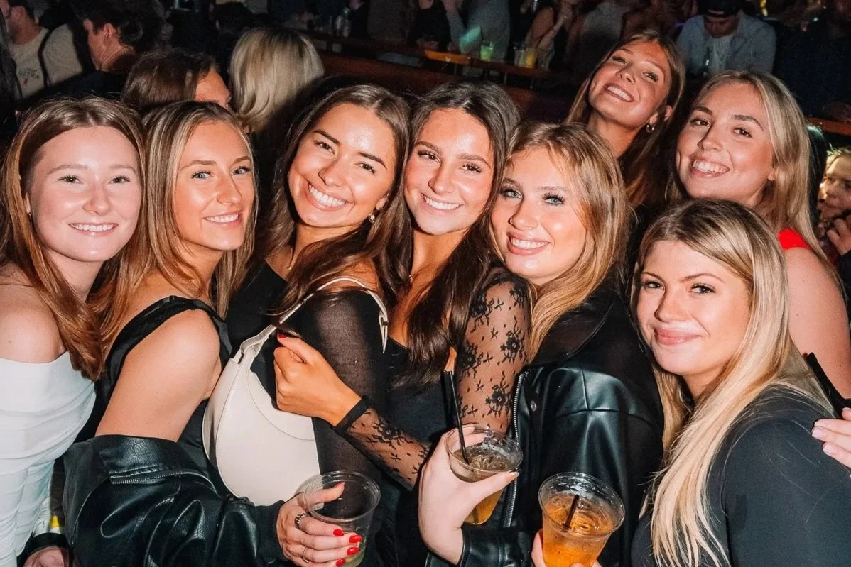 Group of nine young women celebrating at a party, smiling, holding drinks, and dressed casually and stylishly.