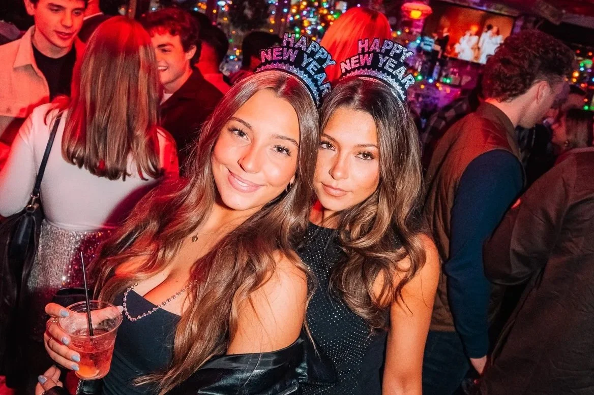 Two young women wearing 'Happy New Year' headbands at a New Year's party, smiling and posing for the camera. They are surrounded by other party attendees in a festive, colorful setting.