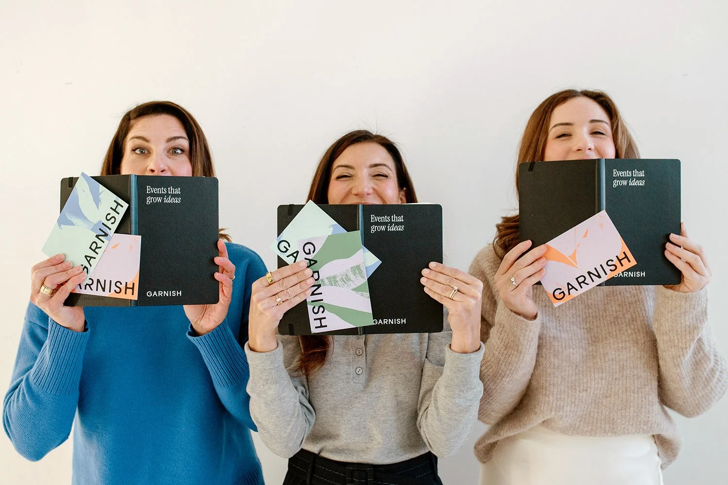 Three women holding black notebooks with GARNISH events notebooks, displaying plant-themed Garnish stickers, smiling behind the notebooks in front of a plain white wall.