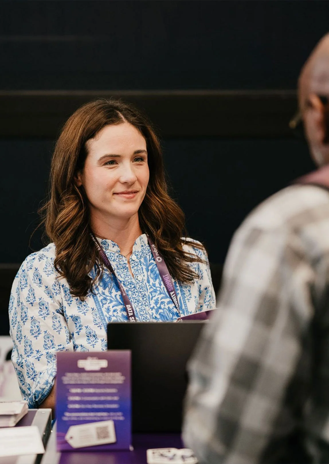 A woman with long brown hair smiling during a conversation at a professional event, wearing a blue and white patterned blouse and a purple lanyard. With informational materials and a laptop in front of her. An out-of-focus person is in foreground.