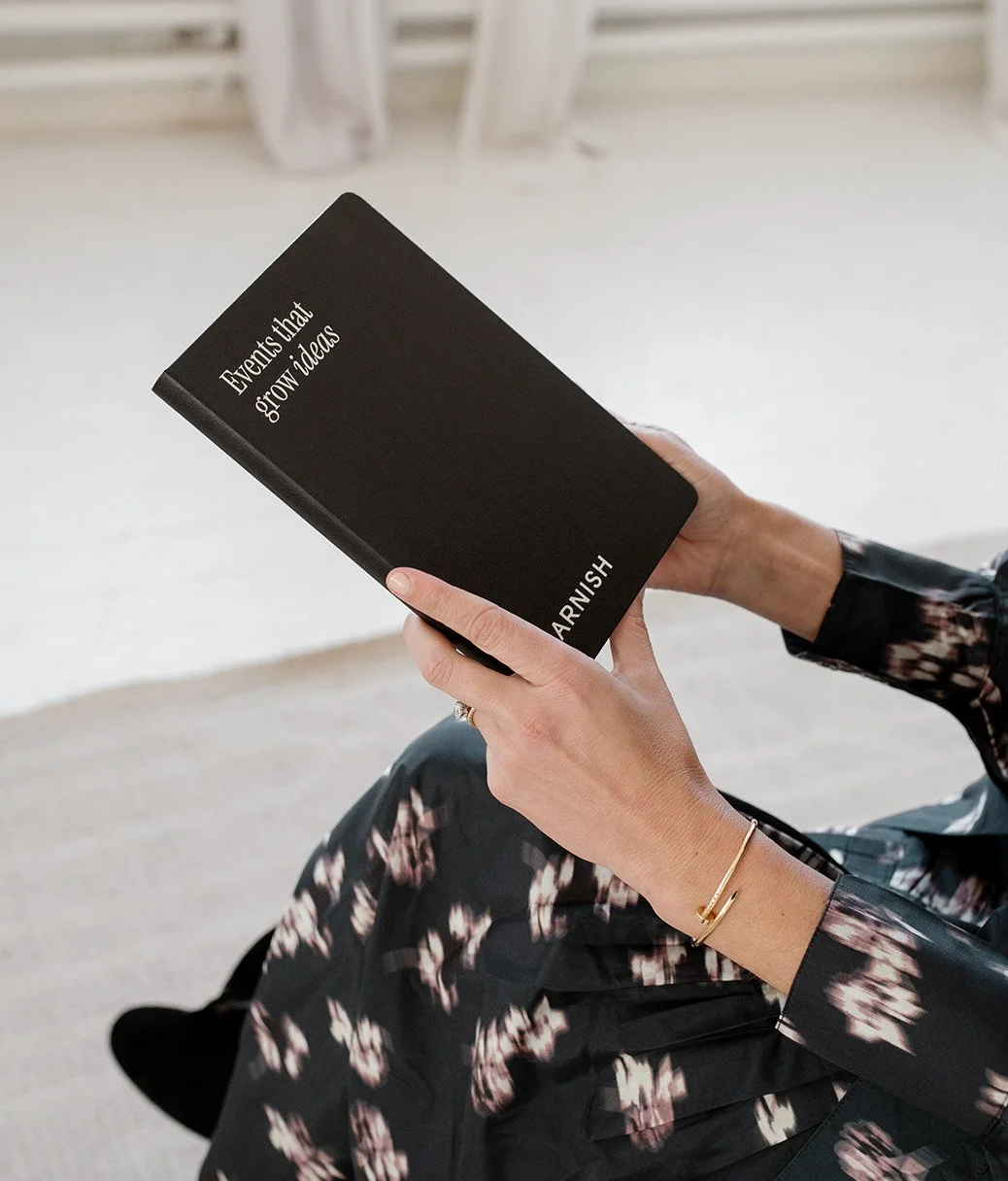 A person holding a black book titled, Garnish "Events that grow ideas", sitting on a light-colored surface, wearing a floral dress, a gold bracelet, and a ring.