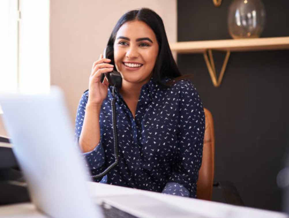 A woman with dark hair and a blue patterned shirt is sitting at a desk, smiling, and talking on a landline phone.