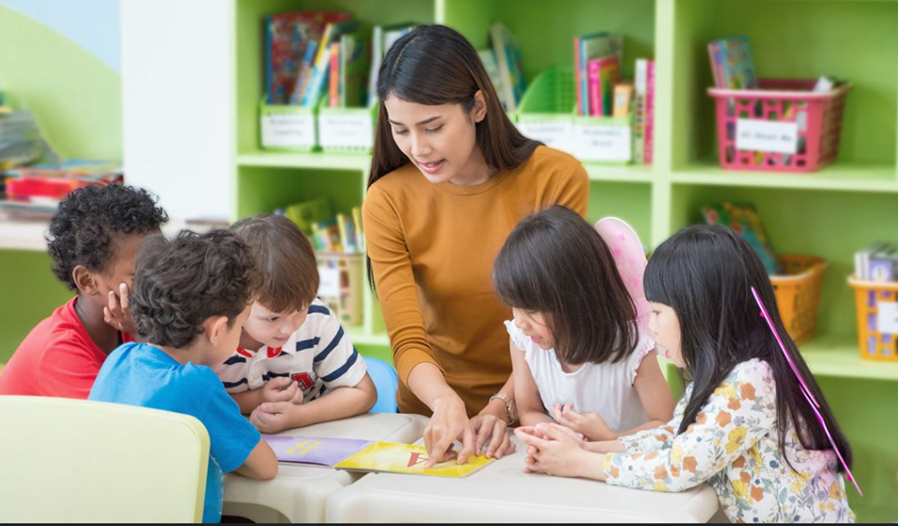 A teacher reading a colorful book to five children in a classroom with green shelves filled with books and baskets.