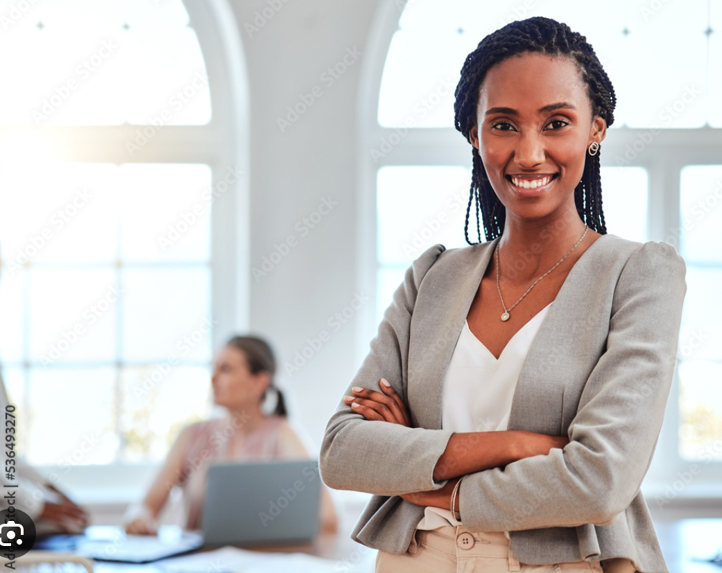 A smiling woman with braided hair in a gray blazer, standing in a bright office with large windows, with a woman working at a desk in the background.