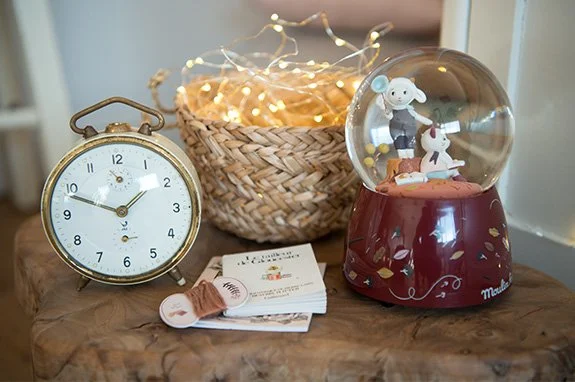 Un reloj despertador dorado, una cesta con luces de cadenas, una bola de nieve con figuras de un niño y un perro, y algunos papeles y cinta en una mesa de madera.