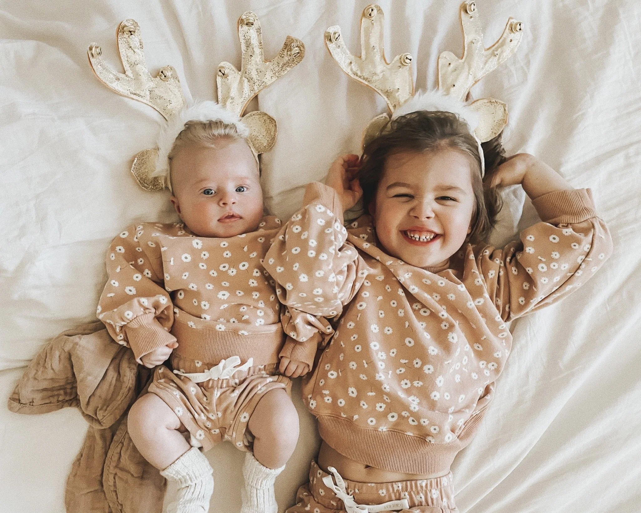Dos niños con orejas de reno, una niña mayor y una bebé, acostados en una cama de pañuelo beige con ropa a juego con estampado de flores blancas, sonriendo y descansando. La niña mayor está sonriendo ampliamente, mostrando sus dientes y levantando las manos detrás de su cabeza.