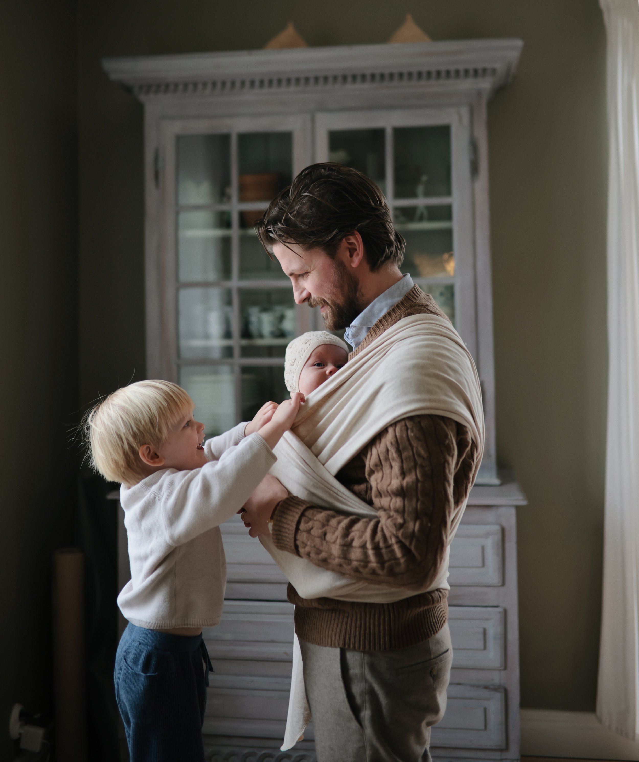 Hombre con barba sosteniendo a un bebé envuelto en un portabebés, mientras un niño pequeño le jala la ropa y los mira con alegría en un interior hogareño.